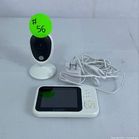 Camera unit with black lens on a white base, handheld display screen, and power adapter cables seen from frontal angle.