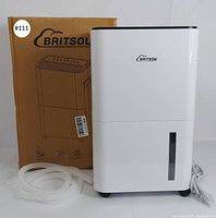 Front view of the BRITSOU dehumidifier showing water level window, included drain hose coiled on the side, and original box with product branding.