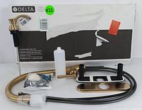 Set of parts and original box with Delta branding, showing gold faucet components, soap dispenser, mounting hardware, and hoses laid out.