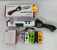 Photo showing the detachable game gun accessory in black and white with blue accent, along with its box, and four Joy-Con controllers in purple, green, yellow, and black.