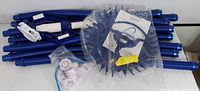 Multiple blue flexible hoses, vacuum head with circular brush, white wheel deflector, small yellow accessory parts, and plastic bag with additional hardware shown.
