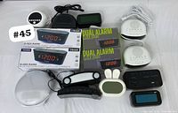 Overview of lot showing multiple digital alarm clocks, Bluetooth radios, and LED time displays in mostly black and white plastic with one pink unit, including dual alarm clock radios in boxes.