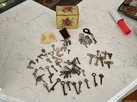 Top-down view showing a variety of antique skeleton keys, clock keys, and an old decorative tin box on a white marble surface.