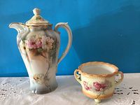 Chocolate pot and sugar bowl side by side on white embroidered cloth, showing floral designs and gold accents.