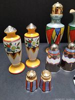 Clear photo showing five salt and pepper sets; mustard yellow floral with handpainted details, black and white geometric, multicolor geometric with silver tops, and berry red geometric. Arrangement highlights Art Deco style and color variety.