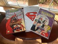 Three items on wooden round table: Overstreet Comic Book Price Guide, sealed Superman comic in plastic with red logo, vintage Blondie comic in protective sleeve.