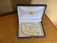 Full view of the freshwater pearl necklace inside its open navy blue presentation box lined with white satin and gold emblem.