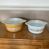 Two Pyrex Butterfly Gold casserole dishes side by side, showing one white with gold design, one gold with white design, both with clear lids.