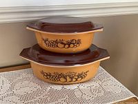 Two vintage Pyrex oval Cinderella casserole dishes stacked, gold color with brown fruit pattern and solid brown lids.