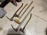 Photo showing all five tools including the yellow sledge hammer, two axes, and two vintage log adzes laid out on a speckled floor.