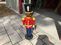 Full front view of the solid wood soldier figure standing outside on a stone-paved surface in front of a building, showing the entire figure including base, body, arms, and head.