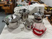 Photo showing all stainless steel cookware items together on a white cloth: large pasta pot with lid, pressure cooker, kettle with cosy, and stock pot.
