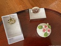 Image of the sterling silver pendant, oval sterling brooch, and porcelain trinket box displayed in white jewelry boxes on a wooden table