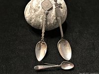 Three items: two collector spoons and a small brooch displayed on a stone for scale and context.
