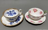 Photo showing two vintage Aynsley fine bone china teacups and saucers, one blue floral and one pink rose floral with a green handle.