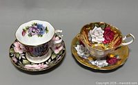 Two vintage fine bone china teacup and saucer sets; one with pink and purple floral pattern and gold trims, the other with gold background and raised floral roses pattern.