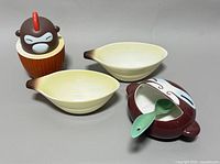 Four ceramic pieces: two oval bowls, monkey head container, teapot-shaped dish with spoon placed on gray surface.
