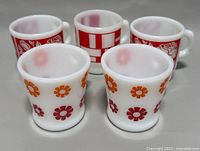 Five milk glass coffee mugs arranged in two rows showing floral and geometric designs.