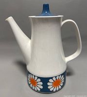 Front view of the white ceramic coffee pot with long spout, curved handle, blue lid, and daisy floral pattern band around the base.