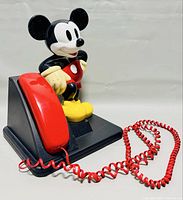 Side view of the vintage Disney Mickey Mouse phone with a bright red handset and coiled cord attached to a black base featuring a standing Mickey figure.