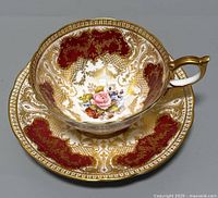 Front-angle view of teacup seated on saucer showing interior floral motif and gilt pattern over red panels