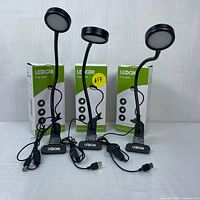 Three LEDGIE clip lights standing upright with boxes behind them, USB cords coiled in front, lights turned off.