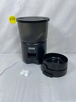 Black automatic pet feeder with 3L food container, digital control panel, detachable feeding bowl, and desiccant pack visible on a white backdrop.