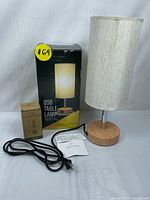 Table lamp with beige fabric shade, wooden and metal base, box, USB LED bulb box, power cord, and instruction manual included.