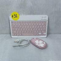 Wireless keyboard with pink keys, white frame and wireless mouse with floral design shown with USB charging cable on reflective silver surface, front view.