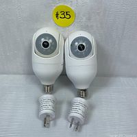 Two white outdoor security cameras shaped like light bulbs with camera lens on the front, shown with attached power socket adapters with US plugs.