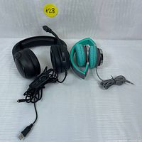 Photo showing both headphones placed on a white backdrop, black gaming headset on the left with attached microphone and USB cable, blue smaller headphones on the right folded with 3.5mm cable coiled.