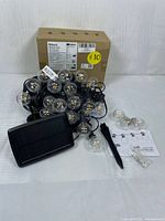 LED solar string lights Model SG08 with black cable, clear globe bulbs, solar panel, ground stake, mounting clips, instruction manual, and original box in background shown from front-view angle.