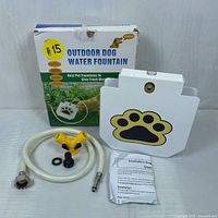 Full view of dog water fountain parts including the fountain unit with paw print, hose, connectors, washers, user manual and box.