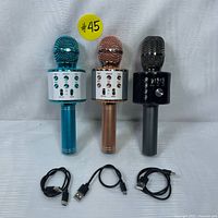 Photo of three wireless Bluetooth microphones in metallic blue, rose gold, and black colors, each standing upright with their USB Type-C charging cables placed in front.