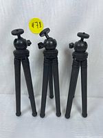 Photo showing three small black mini tripods with adjustable ball heads and textured legs, standing upright against a white fabric background.
