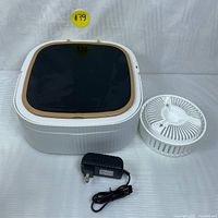 Foldable mini washing machine with lid, agitator basket, and power adapter seen from top and front sides. The machine is white with a dark translucent lid with beige trim, and the body is accordion foldable style. A removable agitator basket and power adapter are shown beside the machine. A flexible drain hose is connected at the back in the front shot.