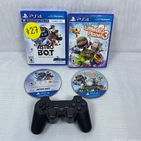 Full view of PS4 wireless controller with Astro Bot and LittleBigPlanet 3 game discs and cases laid on white surface