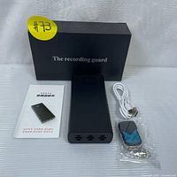 Black rectangular voice recorder on white surface with instruction manual, white USB charging cable, and small black remote control with blue button and metal keyring beside it.