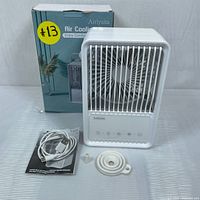 White Airlysita cooling fan with front grille and control panel showing several buttons, placed in front of its retail box. USB Type-C cable, manual, circular mount and small cylindrical mount beside the fan on a striped surface.