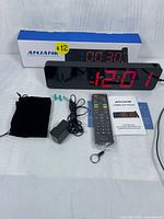Photo showing Anjank 15" LED digital gym timer wall clock powered on displaying time '12:01' and timer '00:30'. Includes remote control, power adapter with cord, mounting screws and anchors, black fabric pouch, user manual, and original box.