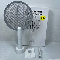 Photo showing the TMACTIME electric fly swatter on its charging stand with the USB cable and user manual in front, along with the original box in the background.