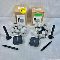 Two sets of white rose solar LED string lights with solar panels and ground stakes displayed next to original product boxes.