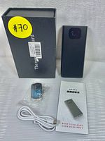 Voice recording blocker device shown with black packaging box, white charging cable, keychain remote, and user manual.