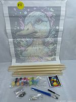 Photo of one paint-by-number canvas rolled out showing graffiti duck design, along with wooden frame pieces, numbered paint pots, brushes, and reference image.
