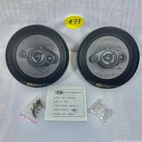 Two round black SAM US speakers with protective grills, mounting screws in plastic bags and test certificate placed on a white surface.