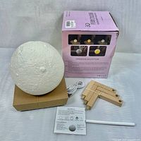 Photo showing the 3D moon lamp, packaging box with instructions, white USB cable, unassembled wooden stand pieces, and user manual, all arranged on a cloth background.