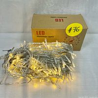 Photo of warm white LED Christmas string lights coiled with clear wiring in front of a brown packaging box labeled 'LED Christmas Lights'. Lights are off in this image.