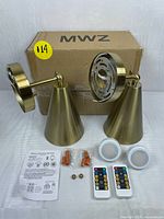 Two gold bathroom light fixtures with mounting plates and conical shades placed in front of MWZ branded box. Accessories including battery-operated LED light bulbs, remote controls, mounting hardware, and instruction sheets arranged neatly on the surface.