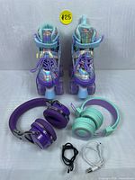 Front view of pair of Nemone adjustable light-up quad roller skates with purple and teal Bluetooth headphones lying in front along with USB cables.