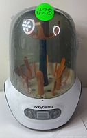 Baby Brezza sterilizer with clear dome cover and orange accessory holders inside, digital control panel on front.